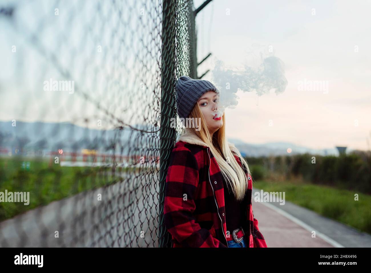 Pretty blonde young Woman with wool hat leaning on the fence and smoking with vaper machine Stock Photo