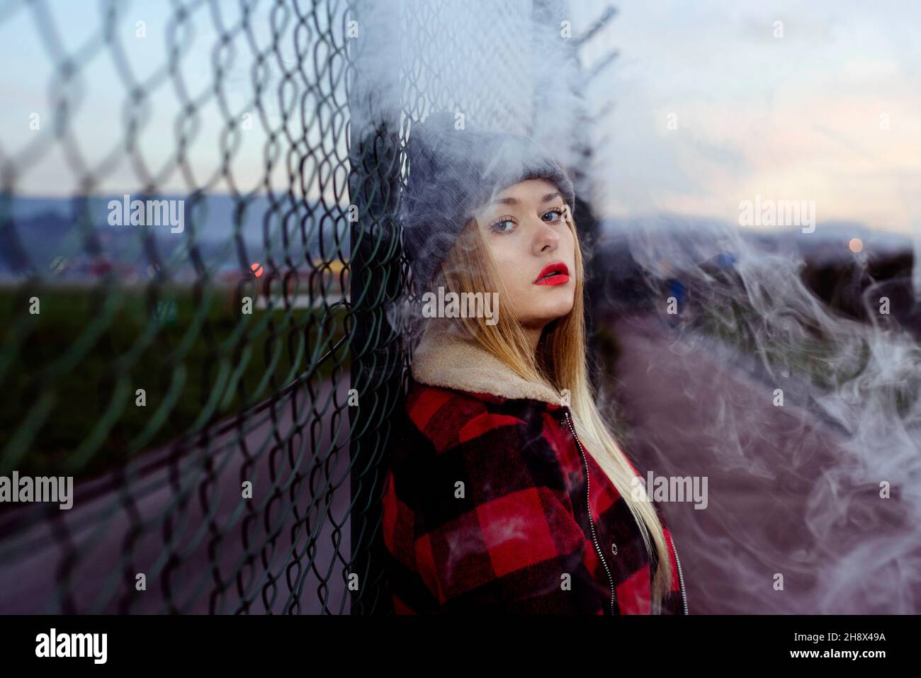 Pretty blonde young Woman with wool hat leaning on the fence and smoking with vaper machine Stock Photo