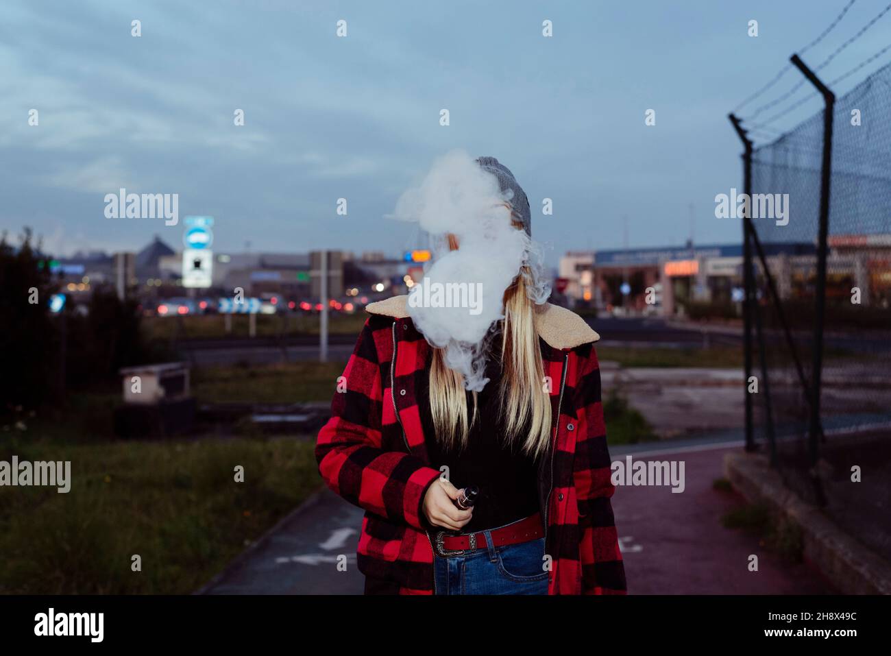 Pretty blonde young Woman with wool hat standing on the street and smoking with vaper machine Stock Photo