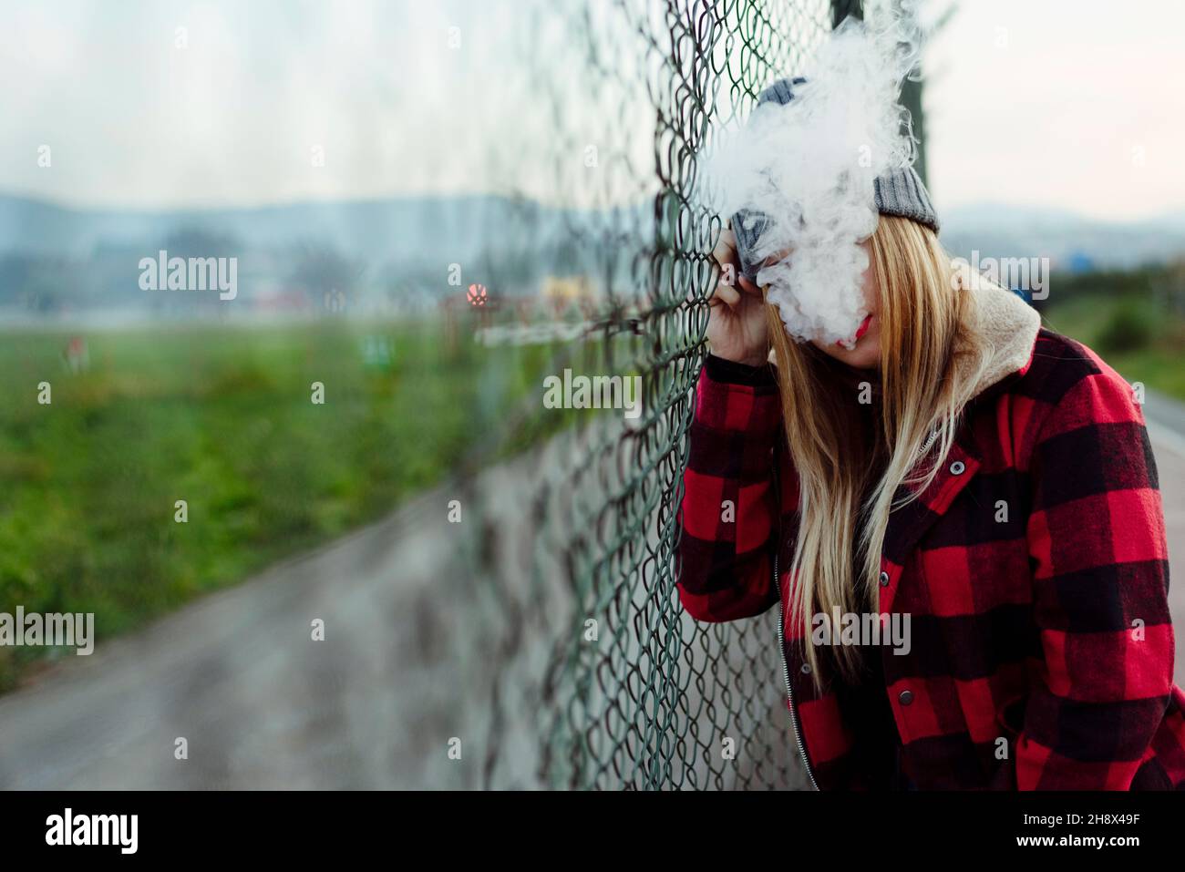 Unrecognizable blonde young Woman with wool hat leaning on the fence and smoking with vaper machine Stock Photo