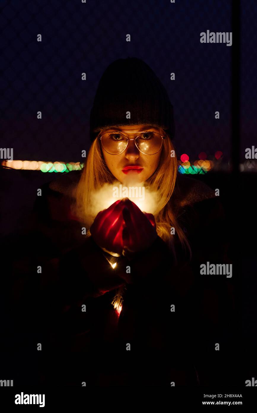 Pretty blonde young Woman with wool hat and glasses holding a light garland of lights looking at the camera on the street Stock Photo