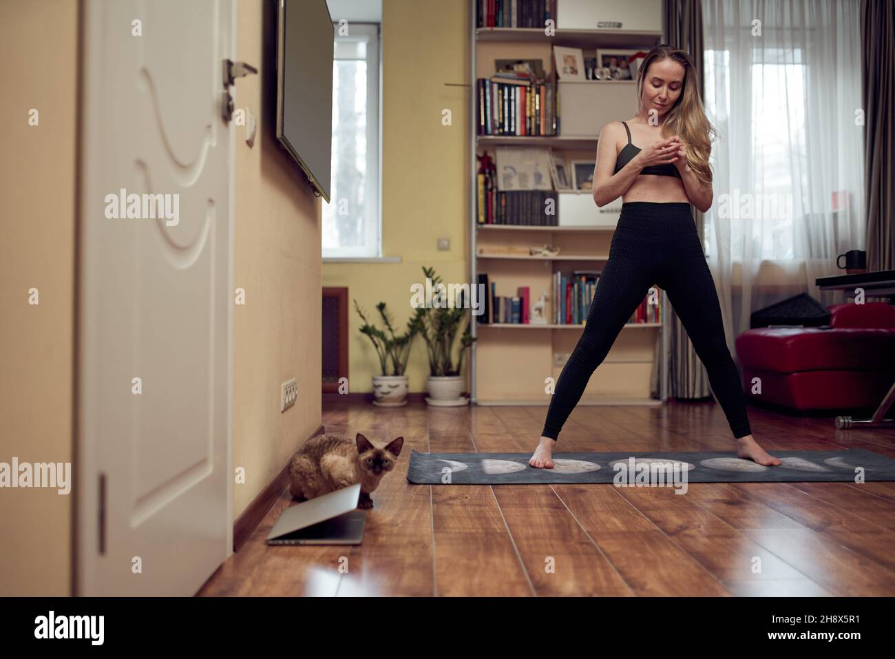 Full body of barefoot female in sportswear standing on mat during yoga session in light living room with laptop and cat Stock Photo