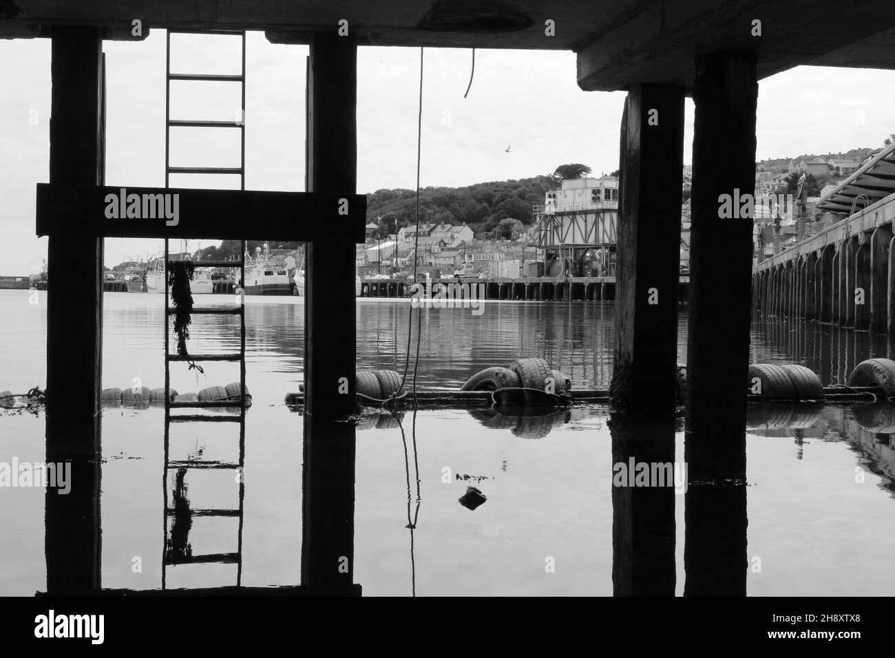 Black and white photo of ladder leading up to a pier, reflected with pillars against the ocean (Newlyn, Cornwall, England). Concept for under the pier Stock Photo