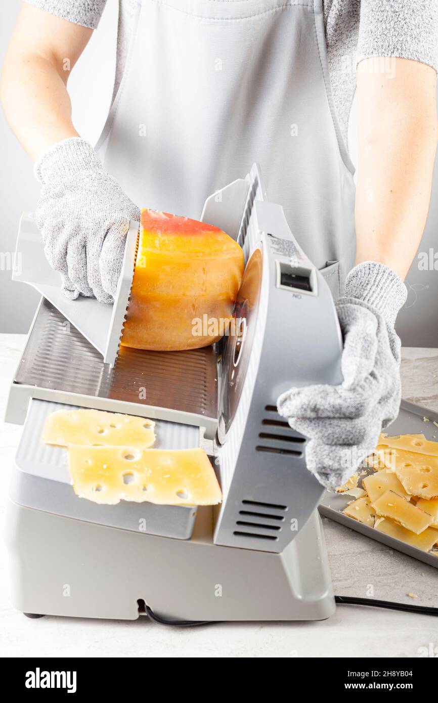 A chef is slicing a large chunk of swiss cheese using an electrical deli slicer. She wears protective cut resistant gloves while operating the machine Stock Photo