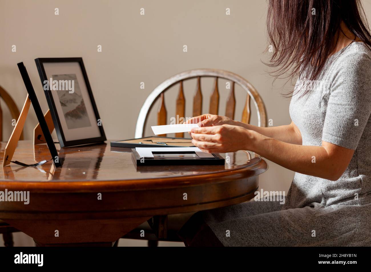 A young caucasian woman is placing a printed photograph into a picture frame with kickstand. There are other frames on the table. Tabletop picture fra Stock Photo