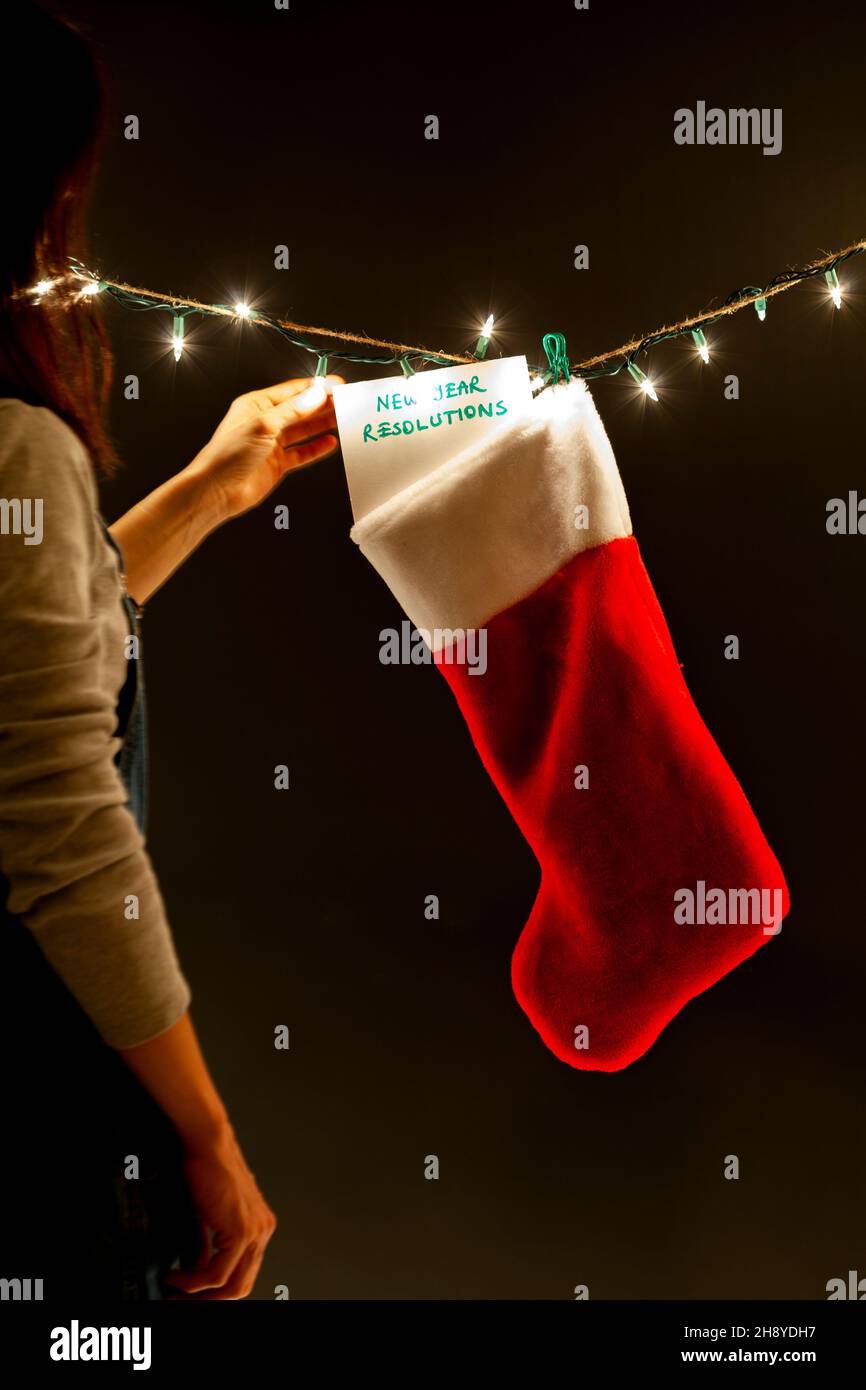 A woman is hanging a notice saying new year resolutions to a a gift stocking on a string of decorative lights. A quirky dark humor concept image for a Stock Photo