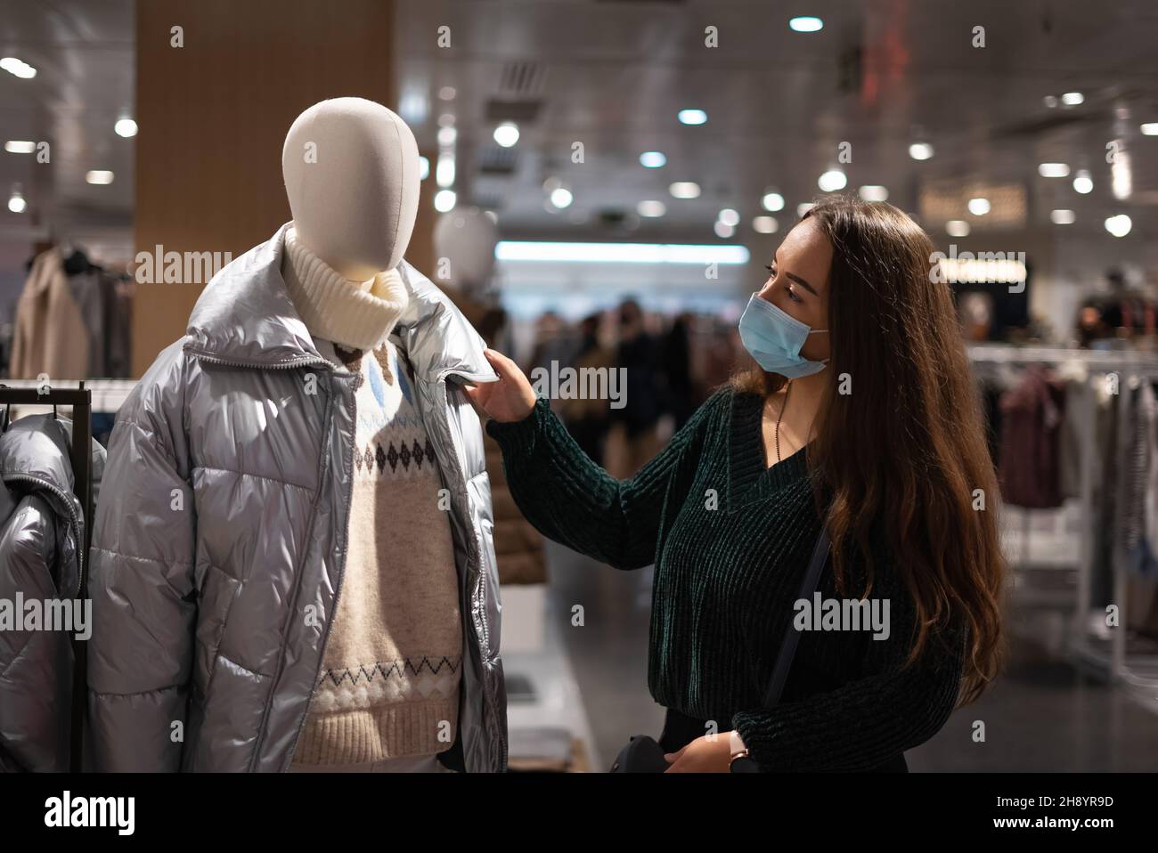 Female customer in mask touching trendy warm coat on mannequin while buying outerwear in modern mall Stock Photo