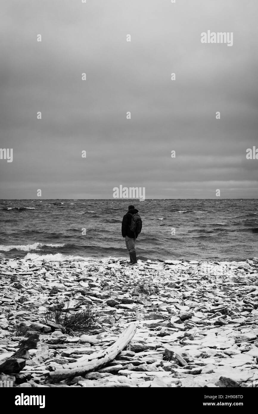 Black and white photo of a man standing at a shoreline with snow on the ground Stock Photo