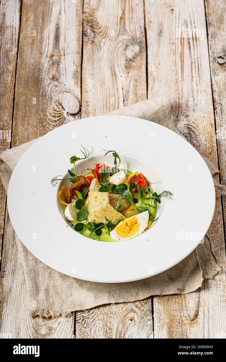 Vertical shot gourmet fish salad. Cod fillet, vegetables, avocado pate and fresh herbs. Restaurant serving. Chef's dish. Healthy and tasty food on a l Stock Photo
