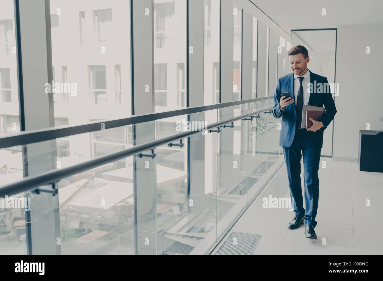 Smiling redhead man office worker in suit using cellphone while standing in modern office Stock Photo