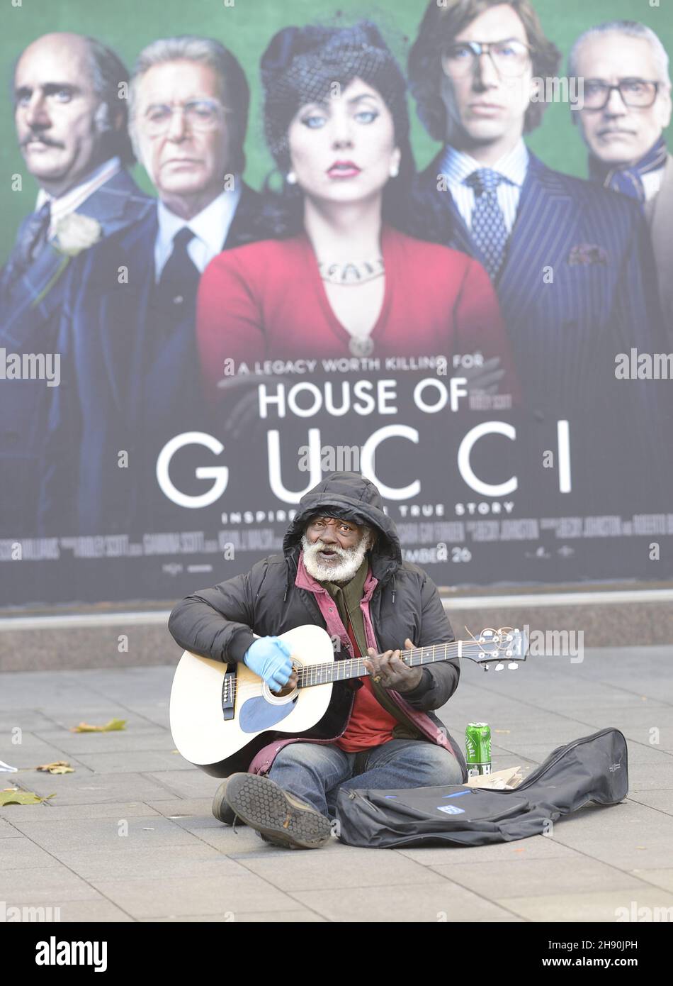 London, England, UK. Elderly black man busking/begging in Leicester Square in fron of afilm poster for House of Gucci - December 2021 Stock Photo