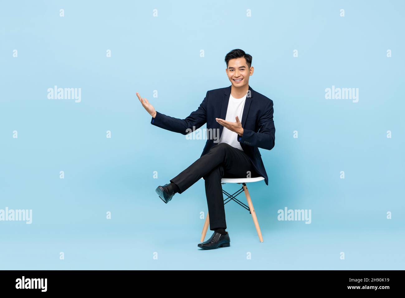 Smiling young Asian man sitting cross legged with hands open to empty space aside in light blue isolated background Stock Photo