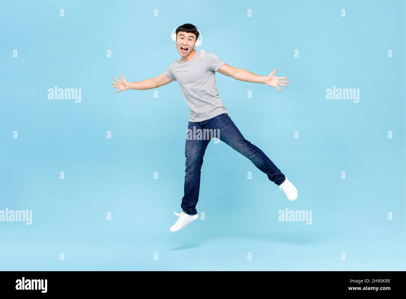 Energetic young Asian man wearing headphones listening to music and jumping on light blue isolated background Stock Photo