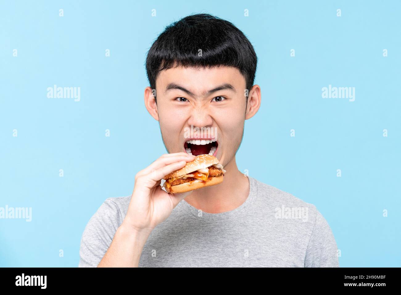 Young hungry Asian man eating hamburger in isolated light blue studio background Stock Photo