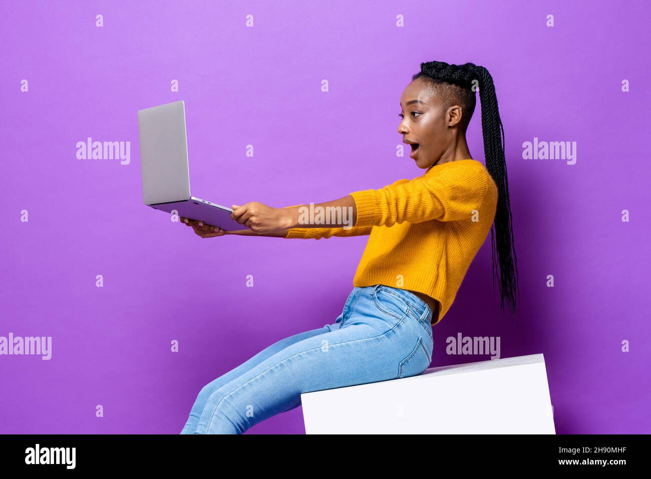 Excited African American woman holding and watching  laptop computer while sitting on stool in isolated purple color studio background Stock Photo