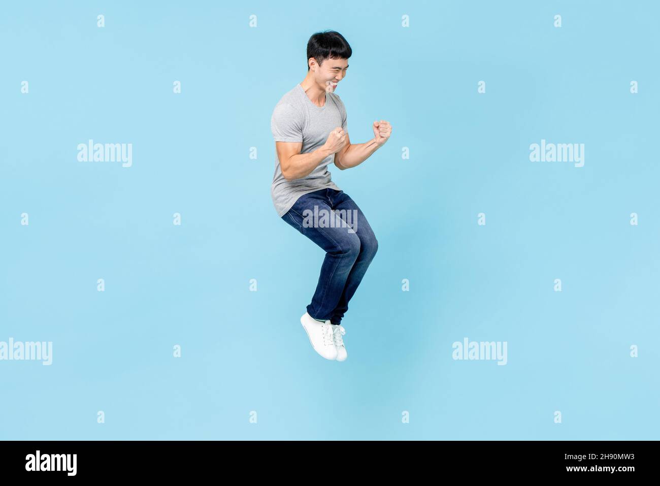 Portrait of smiling young happy Asian man clenching fists and jumping on isolated light blue studio background Stock Photo