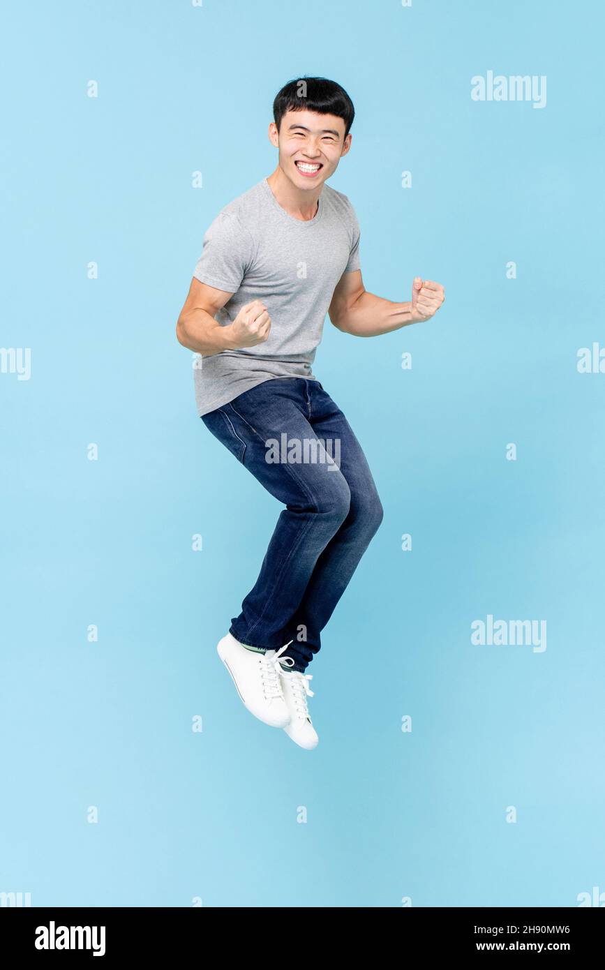 Portrait of smiling young happy Asian man clenching fists and jumping on isolated light blue studio background Stock Photo