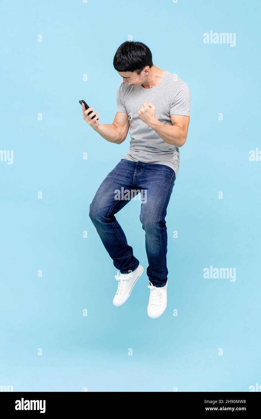 Jumping portrait of smiling young happy healthy Asian man looking at smart phone with fists gesture on isolated light gray studio background Stock Photo