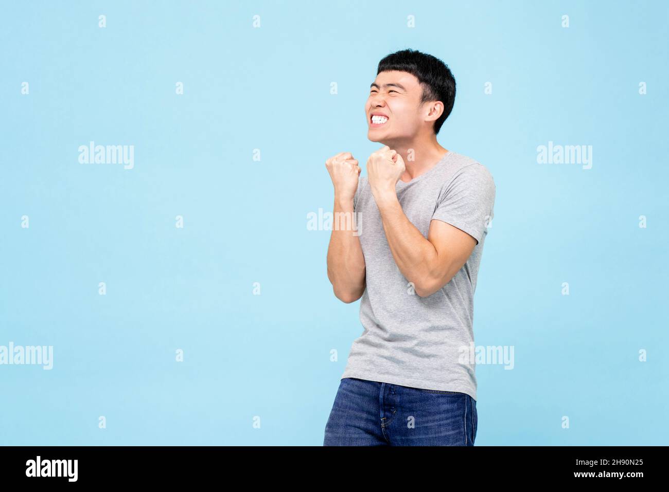 Happy ecstatic young Asian man raising his fists doing yes gesture celebrating success in isolated light blue studio background Stock Photo