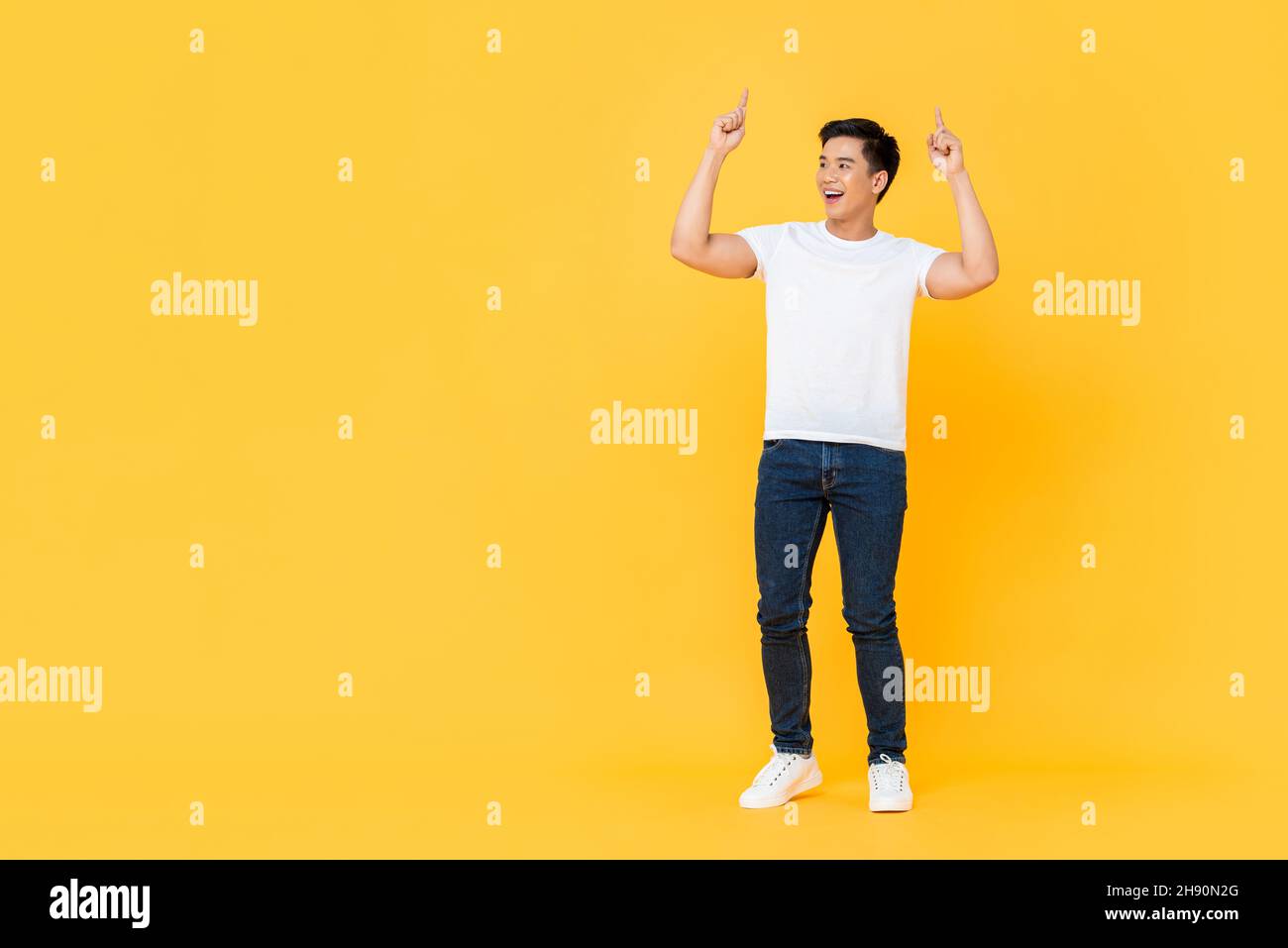 Smiling young handsome Asian man looking at empty space and pointing two fingers upward isolated on yellow studio background Stock Photo