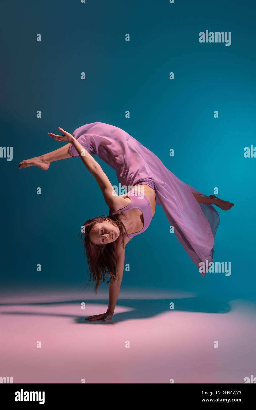 Young adorable flexible contemp dancer in lilac dress dancing isolated on gradient blue white background in neon. Stock Photo