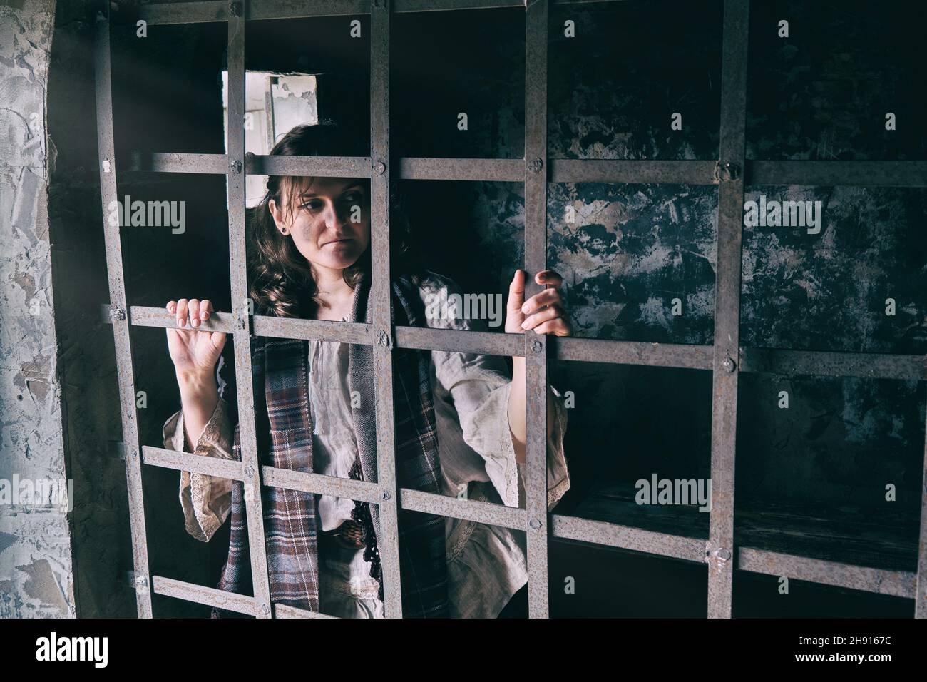 Young brunette woman in vintage clothes behind prison bars, copy space for text Stock Photo