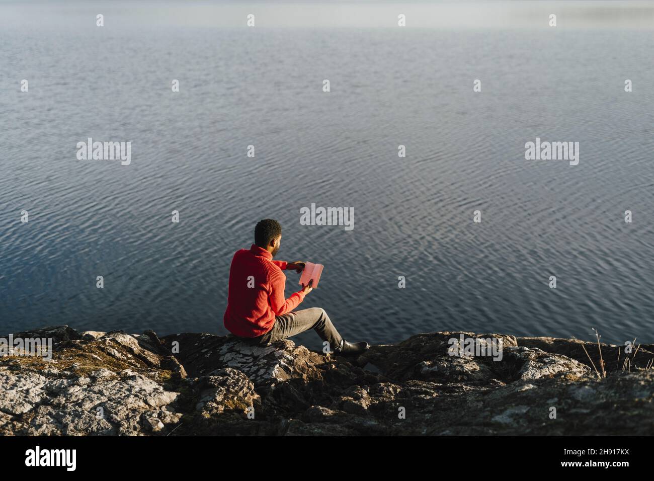 Man reading book while sitting on rock at lakeshore Stock Photo