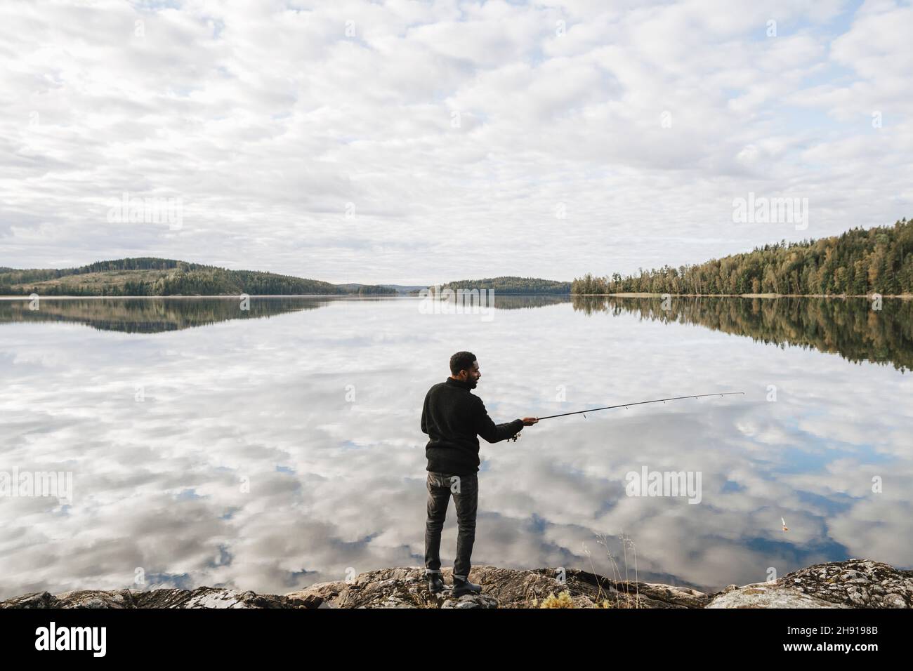 Full length rear view of man fishing in lake with reflection of clouds Stock Photo