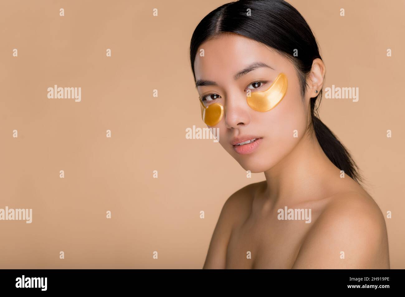Gold eye patches for puffiness, wrinkles and dark circles, skincare concept. Pretty young Asian brunette woman with perfect skin applied gold collagen eye patches,standing on isolated beige background Stock Photo