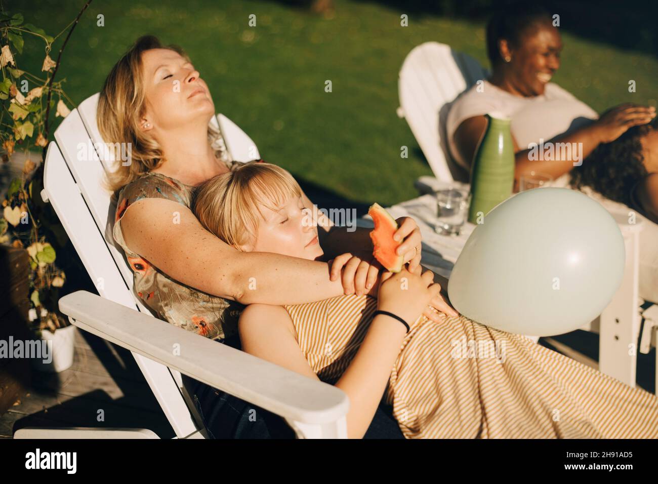 Mother relaxing with daughter in backyard on deck chair during sunny day Stock Photo