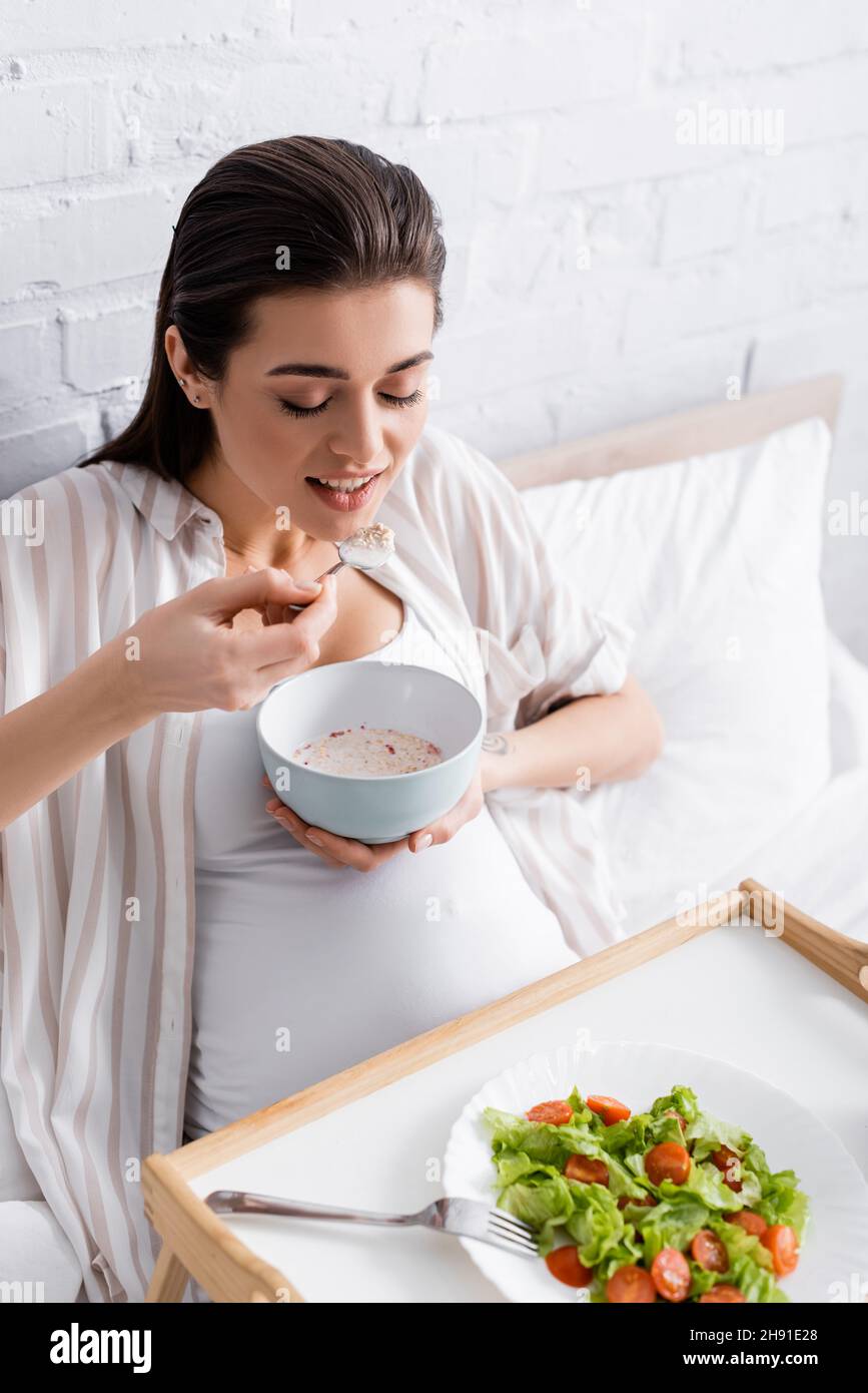 happy pregnant woman eating oatmeal near tray with salad Stock Photo