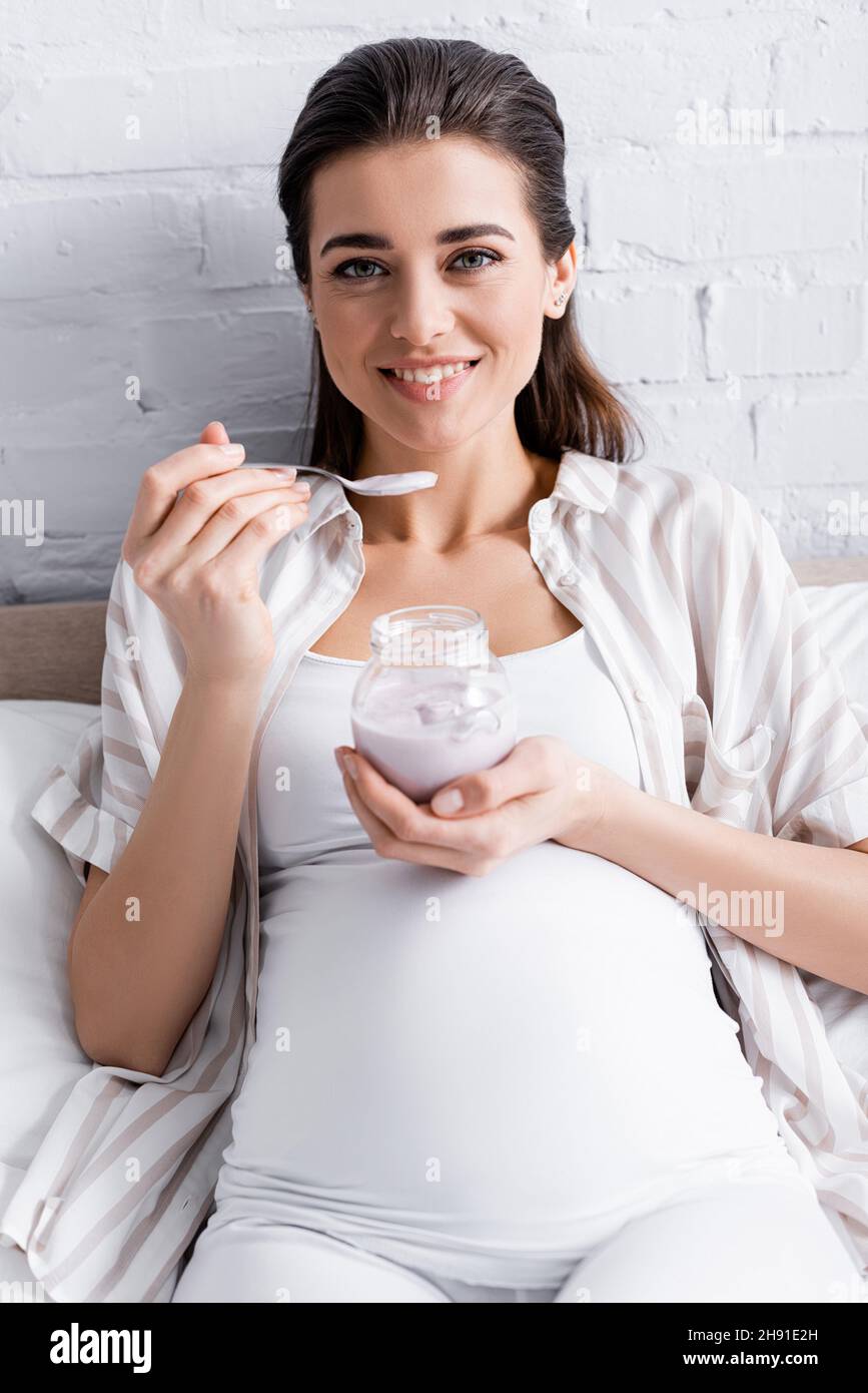 cheerful pregnant woman holding glass jar with yogurt while sitting on