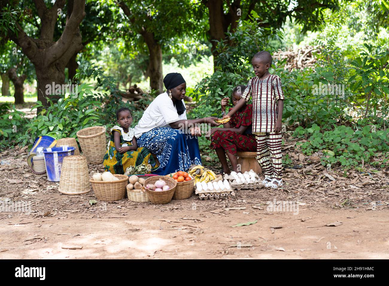 Cute little black boy buying a banana from a group of West African street market vendors sitting under some large mango trees by the roadside Stock Photo