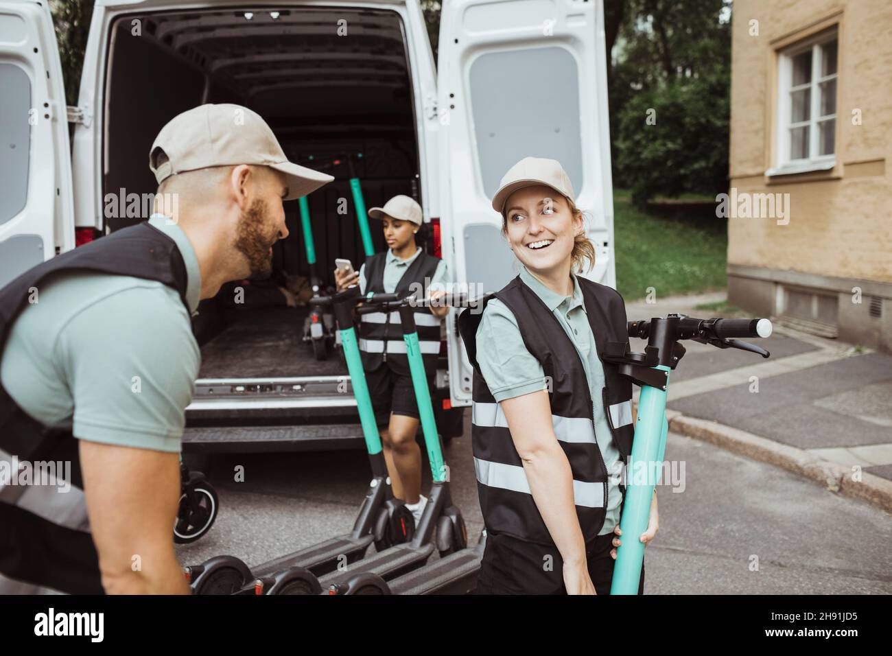 Smiling delivery woman looking at male coworker while loading push scooter in van Stock Photo