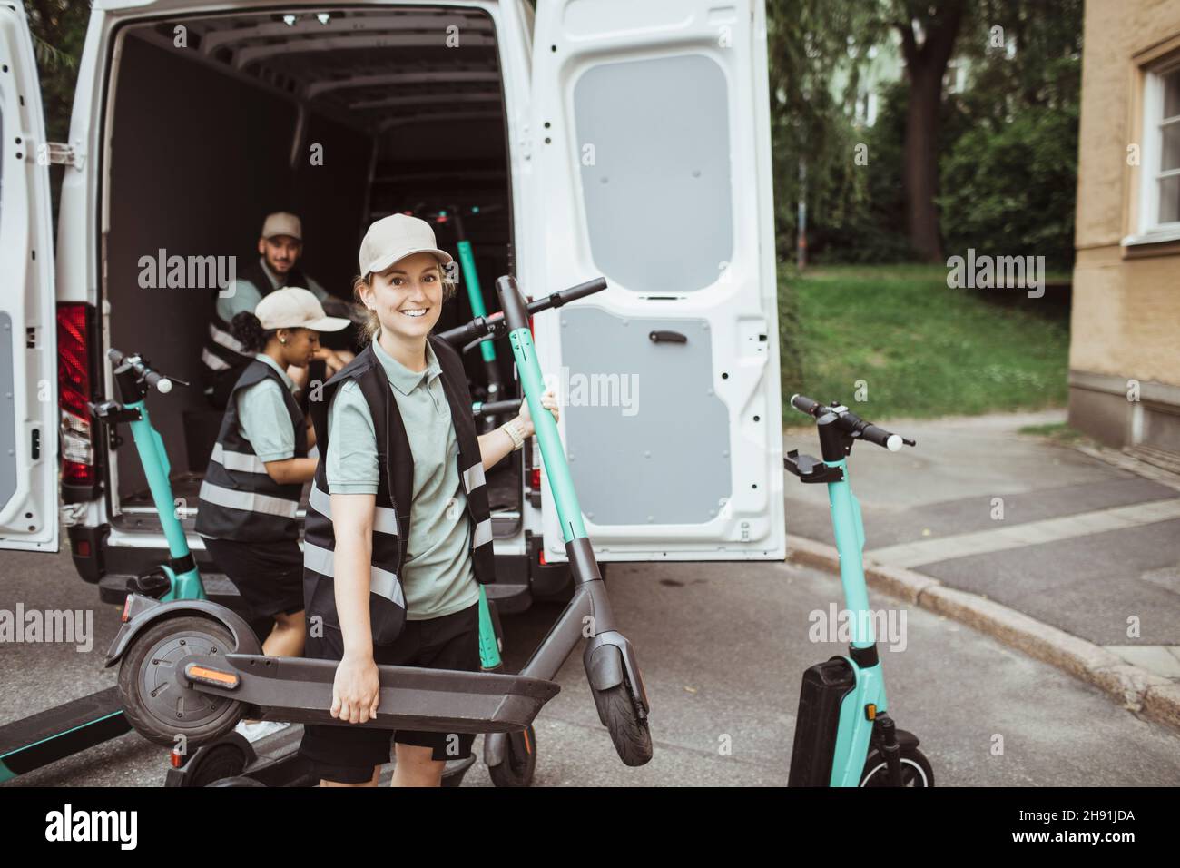 Young deliver woman carrying push scooter while helping male and female coworkers loading van Stock Photo