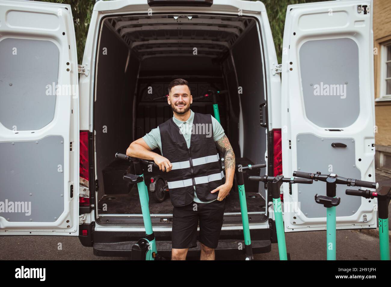 Portrait of smiling male blue-collar worker leaning on push scooter against delivery van Stock Photo