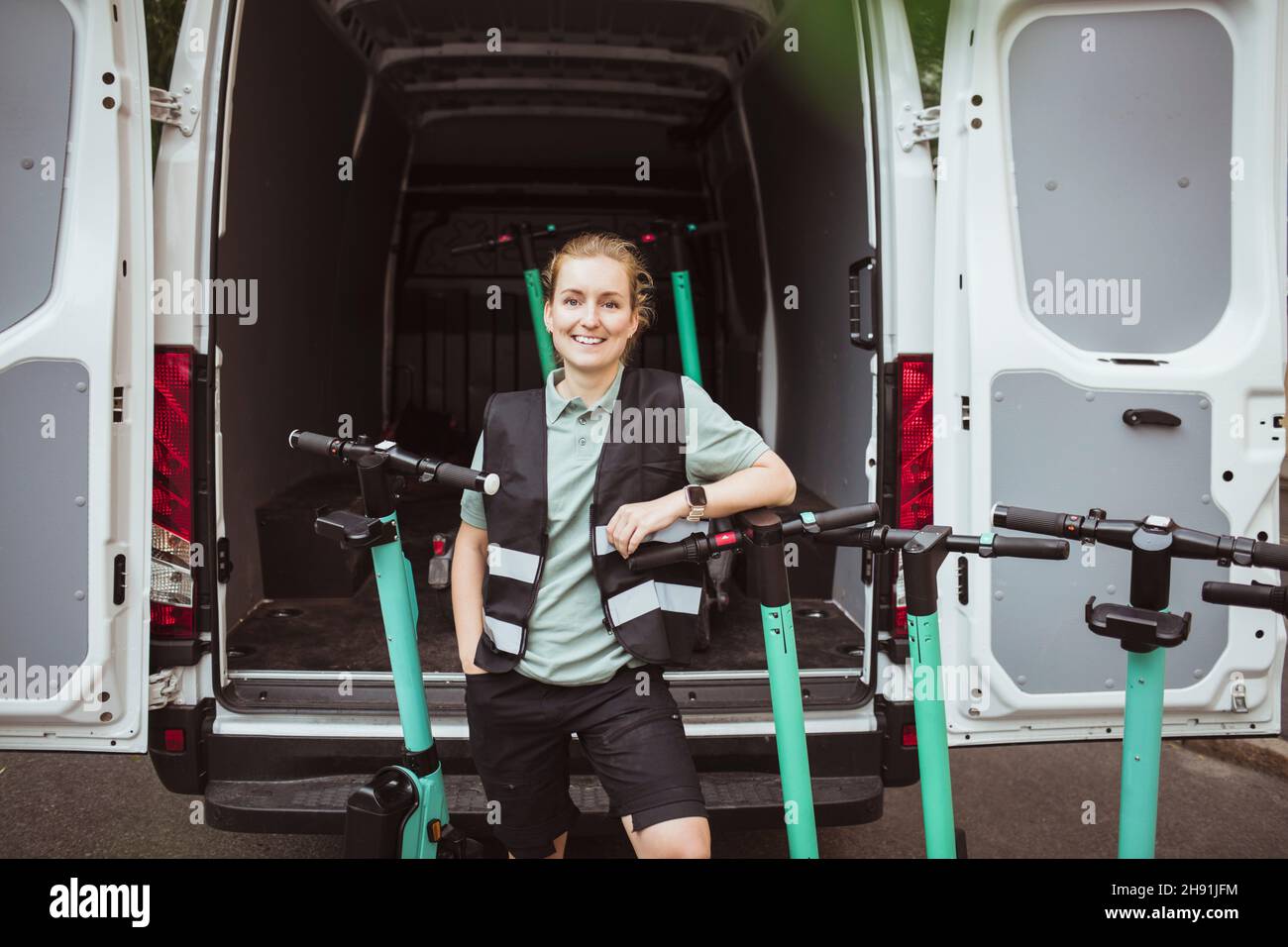 Portrait of smiling female blue-collar worker with push scooters against delivery van Stock Photo