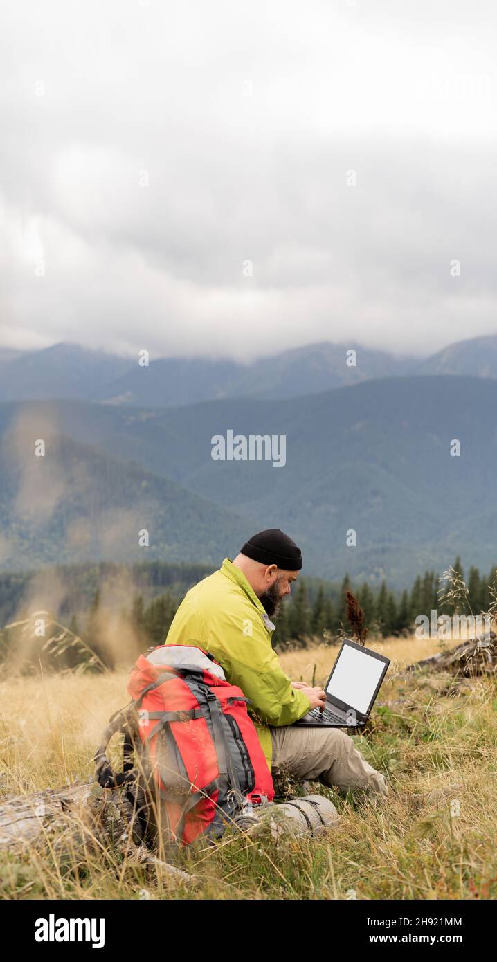 a bearded Caucasian man with a laptop sits on a fallen tree in the mountains with a beautiful view of the valley. Stock Photo