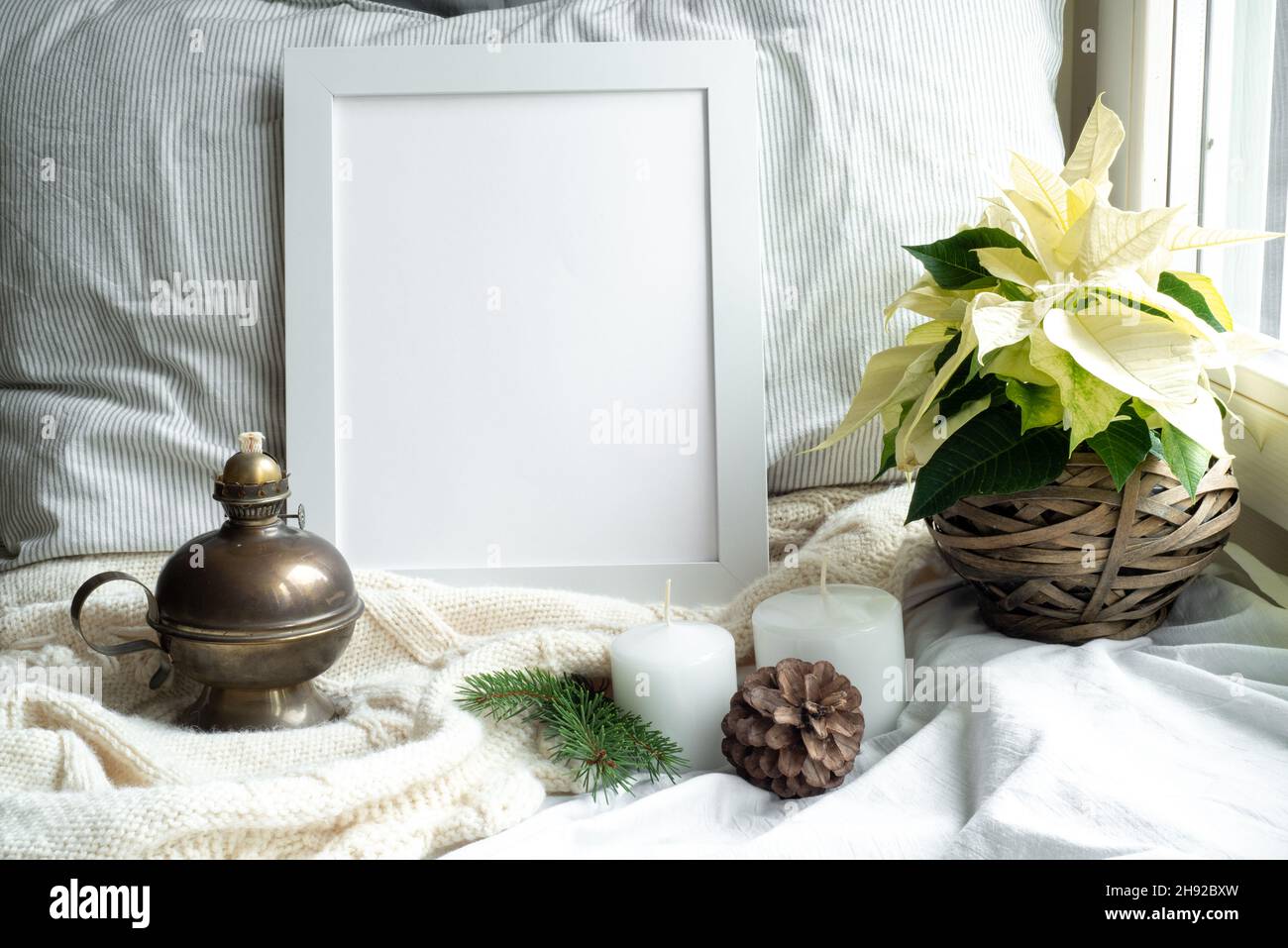 Cozy winter home interior scene.Christmas composition near the window.Blank white pictur frame mockup, white poinsettia, oil lamp, candles, gift box.M Stock Photo