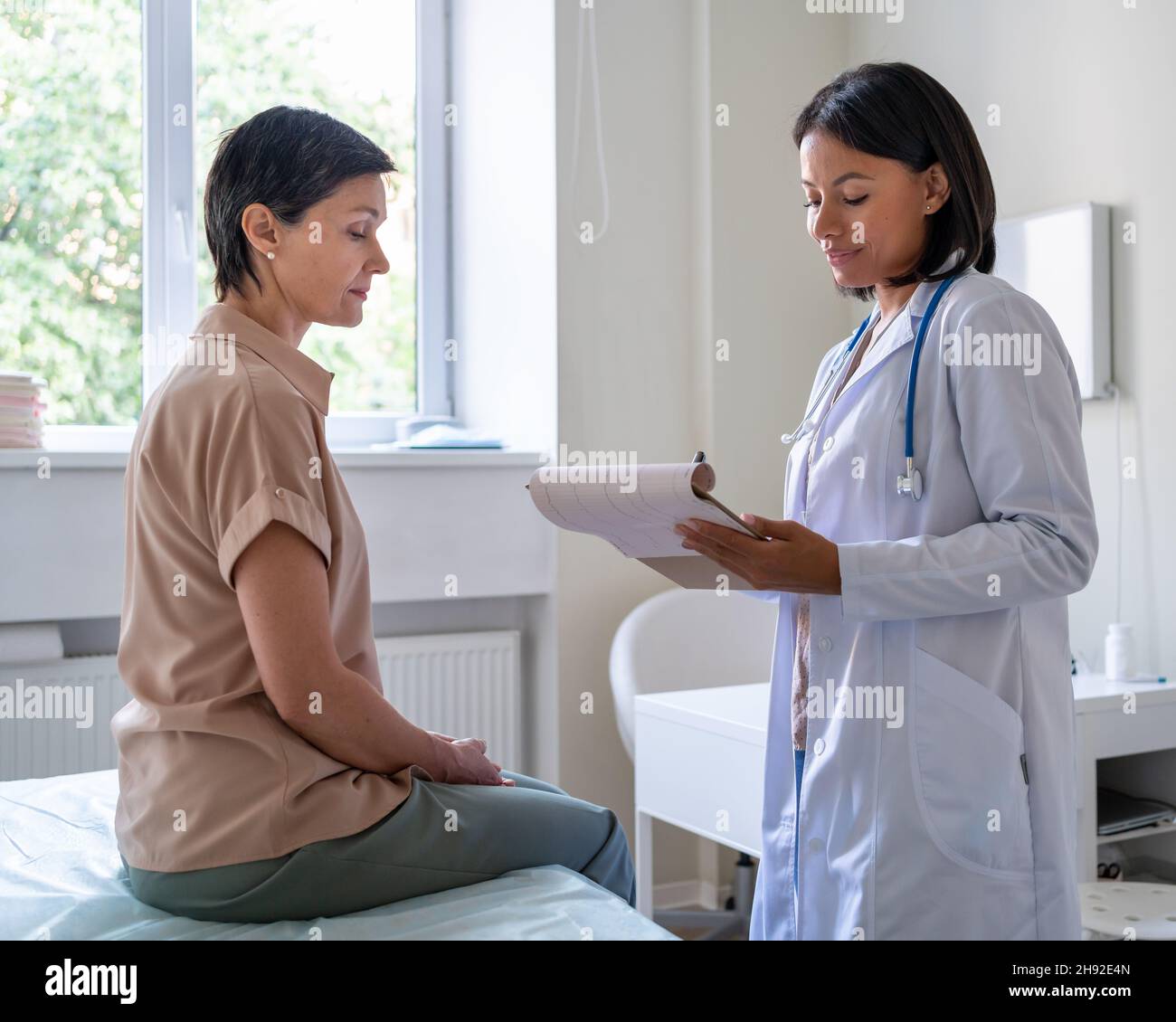 Doctor giving prescriptions on clipboard to middleaged patient sitting on examination couch