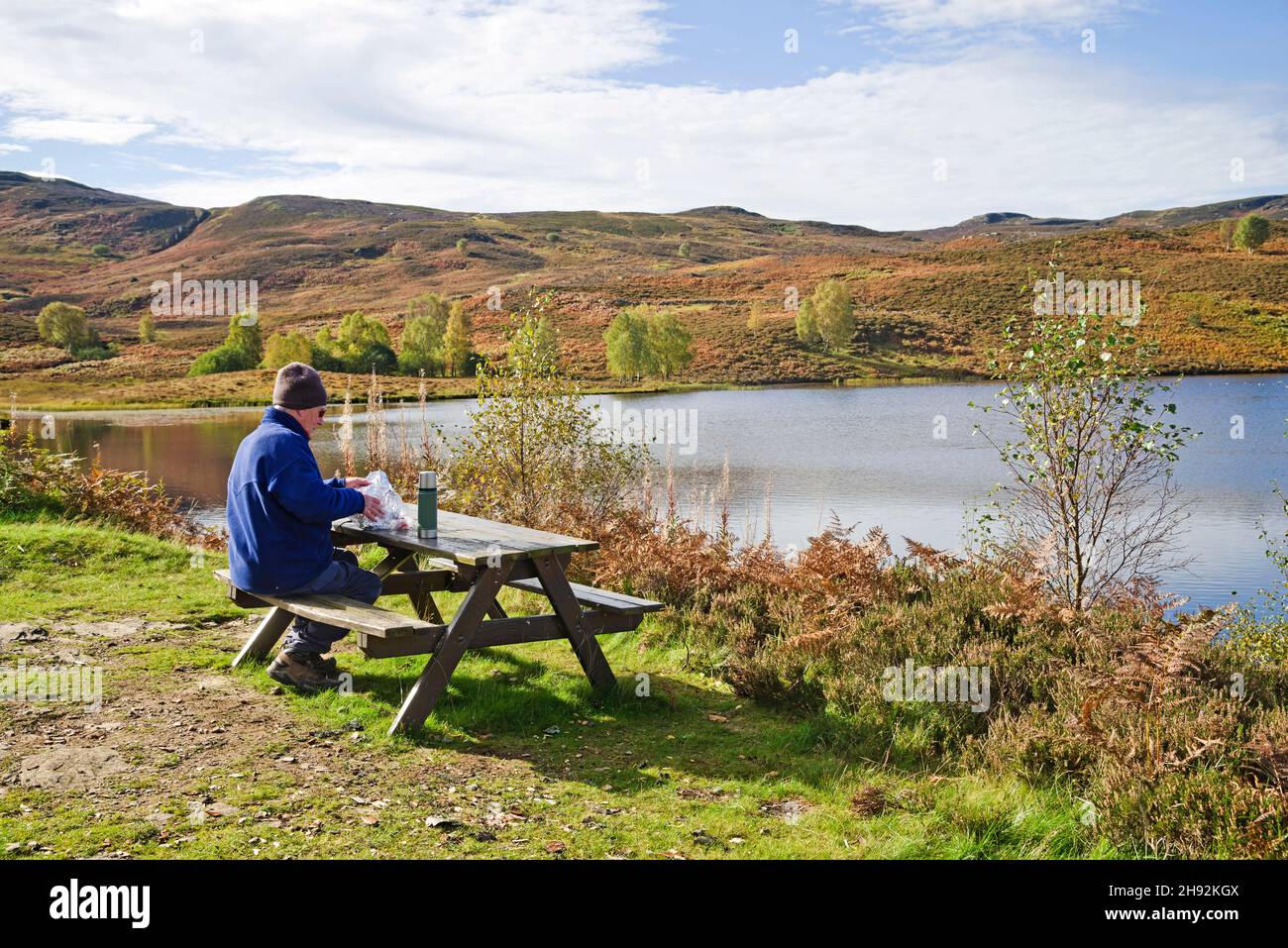 Walker having lunch at picnic table by small moorland hill loch on sunny autumn day, Tayside sporting estate, Highland Perthshire, Scotland UK Stock Photo