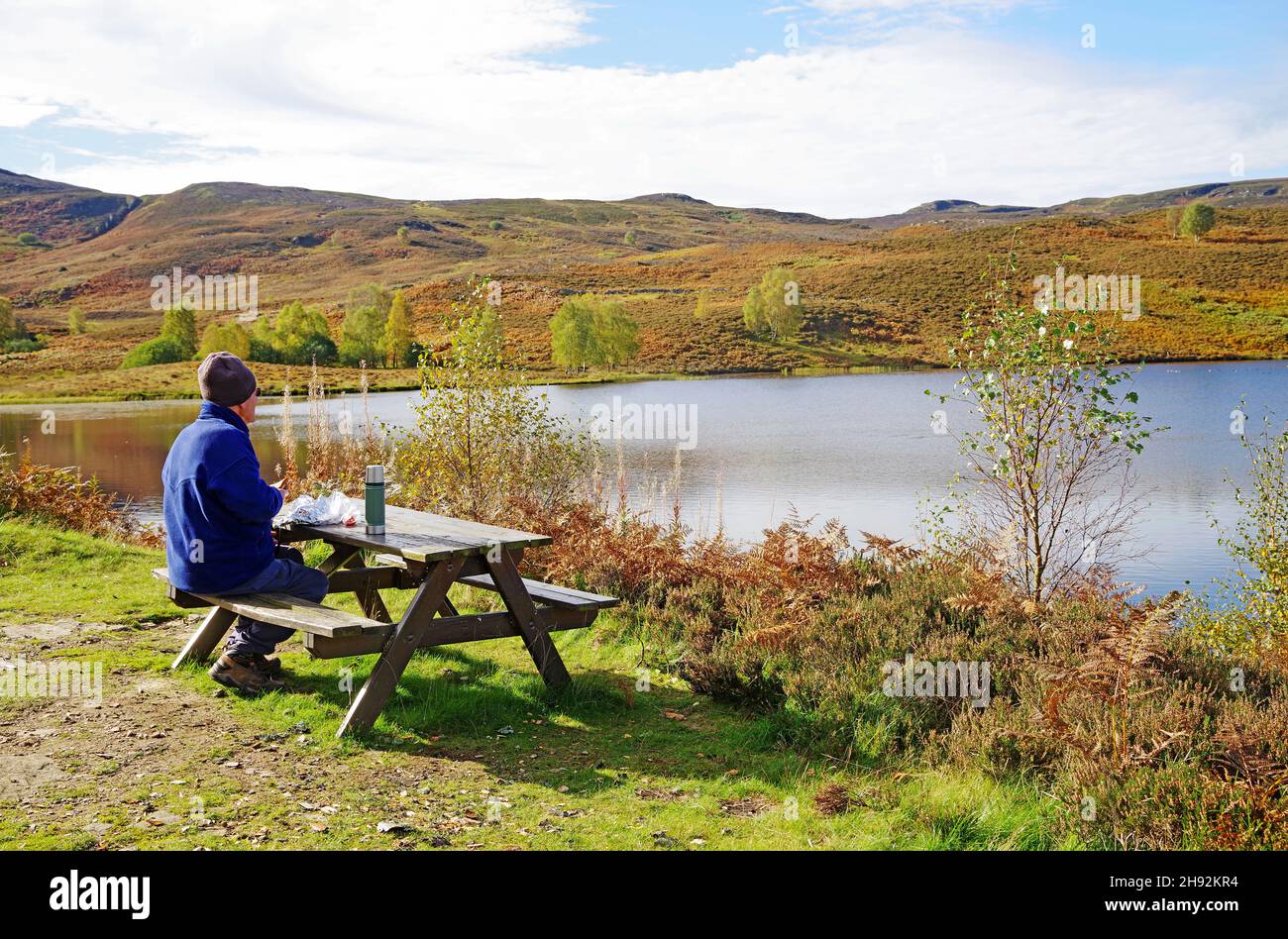 Walker having lunch at picnic table by small moorland hill loch on sunny autumn day, Tayside sporting estate, Highland Perthshire, Scotland UK Stock Photo
