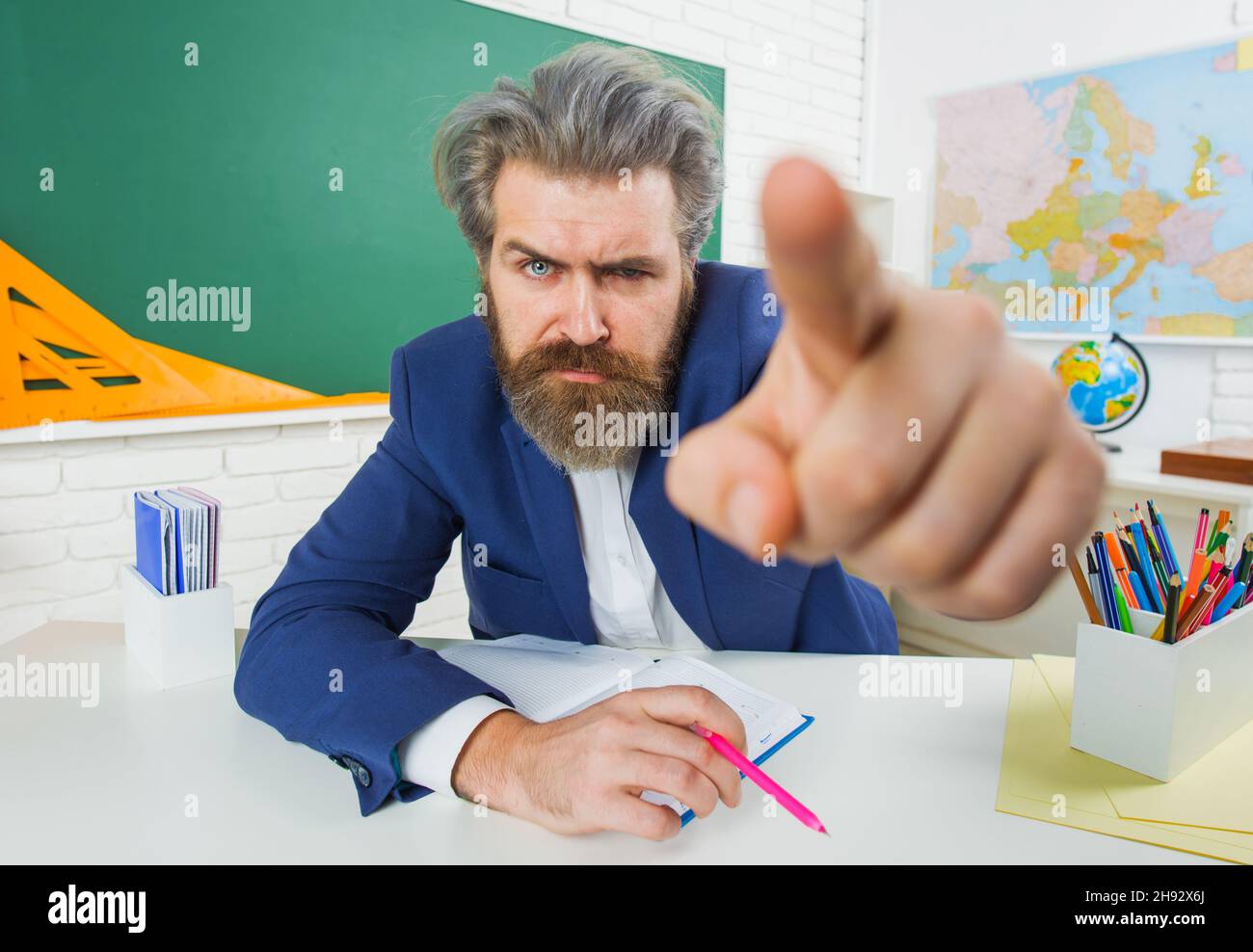 Angry male teacher in classroom pointing finger. Serious bearded man in suit sitting at table in class. Stock Photo