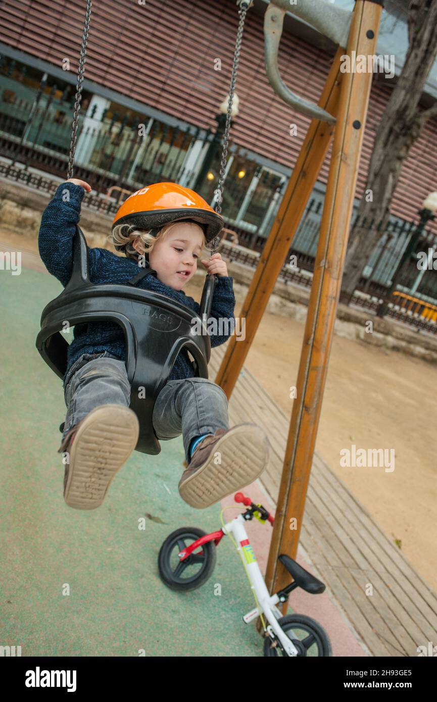 A small boy (ca. 2 1/2 years old) enjoys a ride on a swing in a public park. Resting against the swing's frame is his   pedal-less learner bicycle. Stock Photo