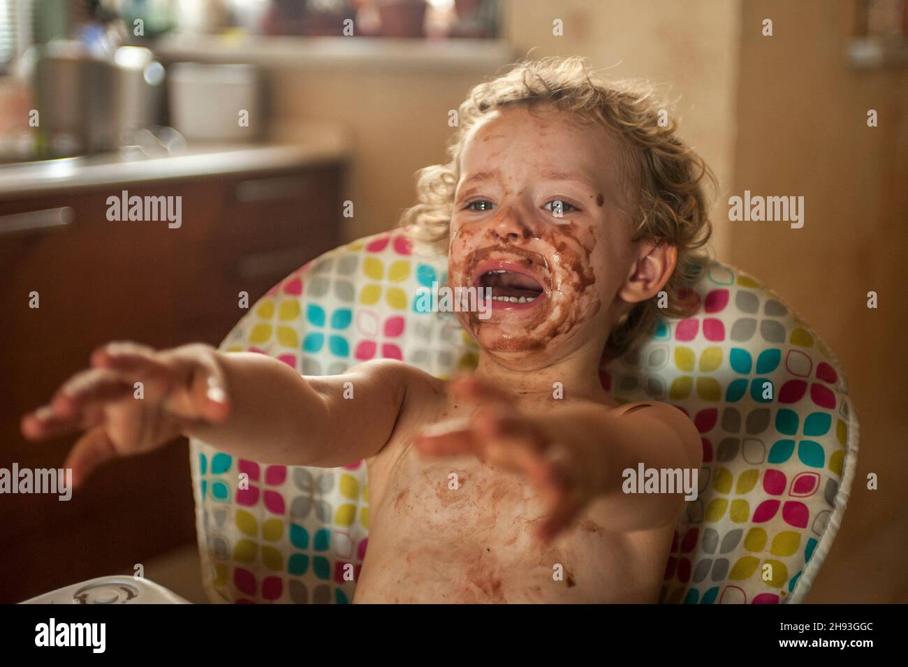A baby girl (age 2 1/2 years) screams and cries while sitting on a high chair and covered in chocolate ice-cream. Stock Photo