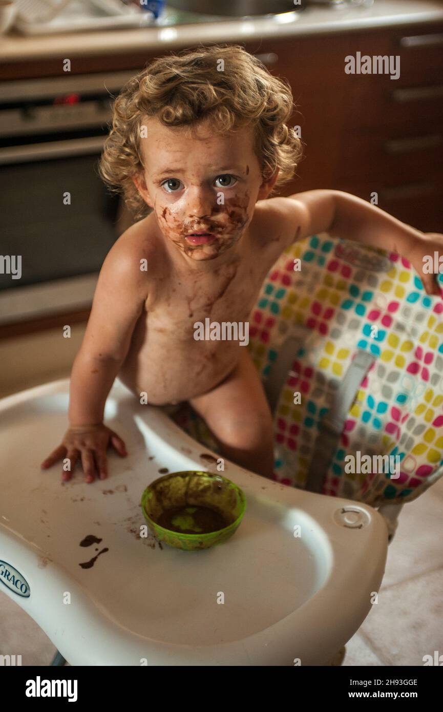 A baby girl (age 2 1/2 years) climbs off a high chair after eating chocolate ice-cream, in which she is covered. Stock Photo