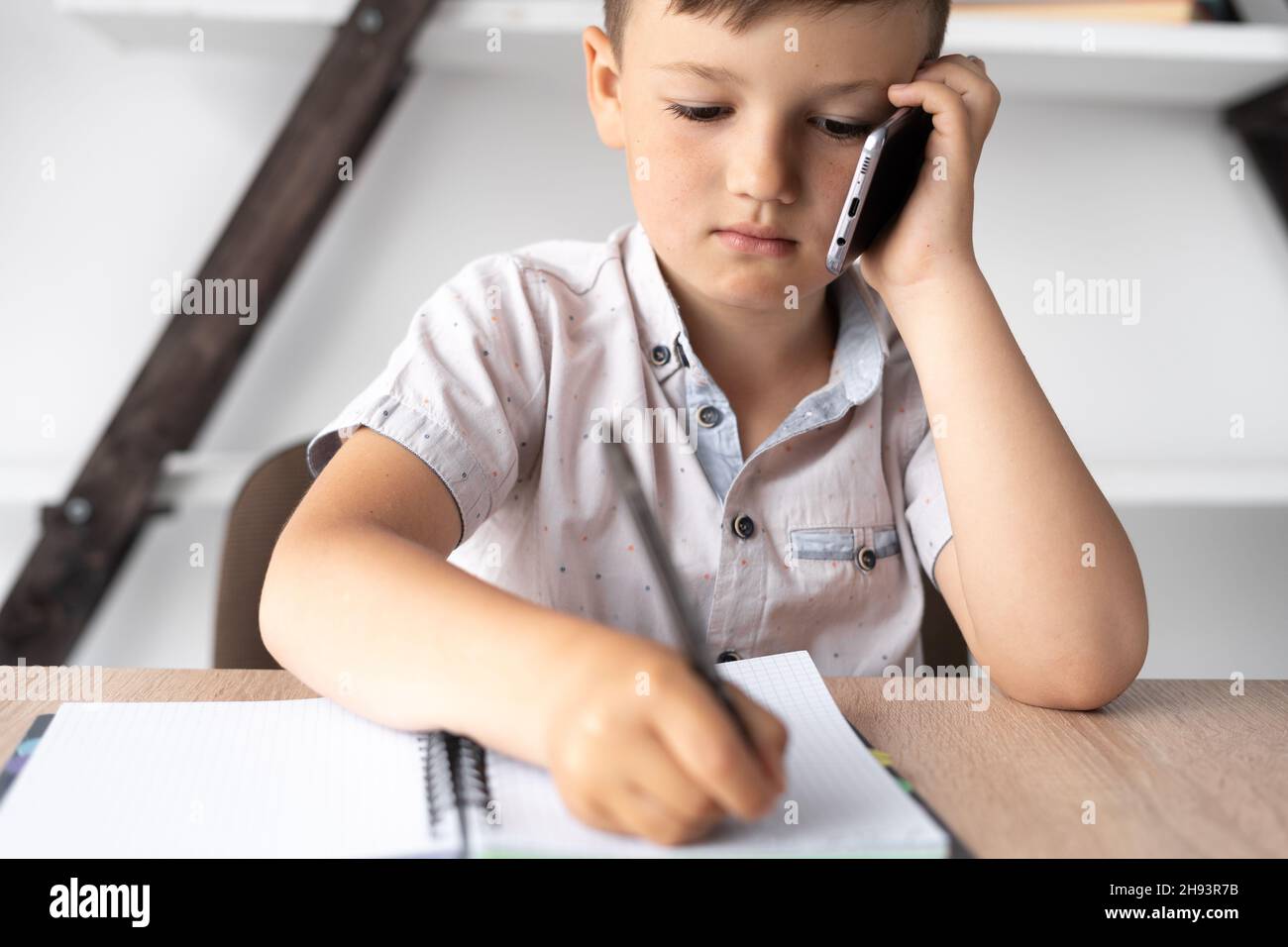 close-up of a little student talking on a cell phone. A boy sits at a desk, talks on a smartphone and makes notes in a notebook. Technology and Stock Photo