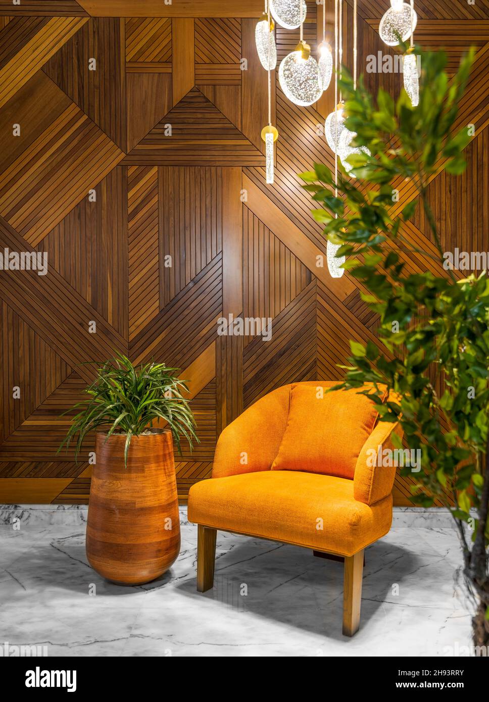 Modern orange armchair, tall wooden planter, and contemporary glass chandelier, in a hall with decorated wood cladding wall, and white marble floor, framed by green bushes Stock Photo
