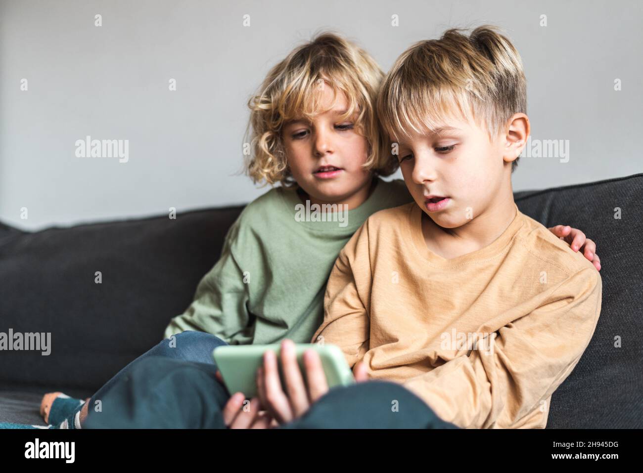 Low angle of calm little sibling with blond hair in casual outfits watching interesting video on smartphone. while sitting together on comfortable sofa at home Stock Photo