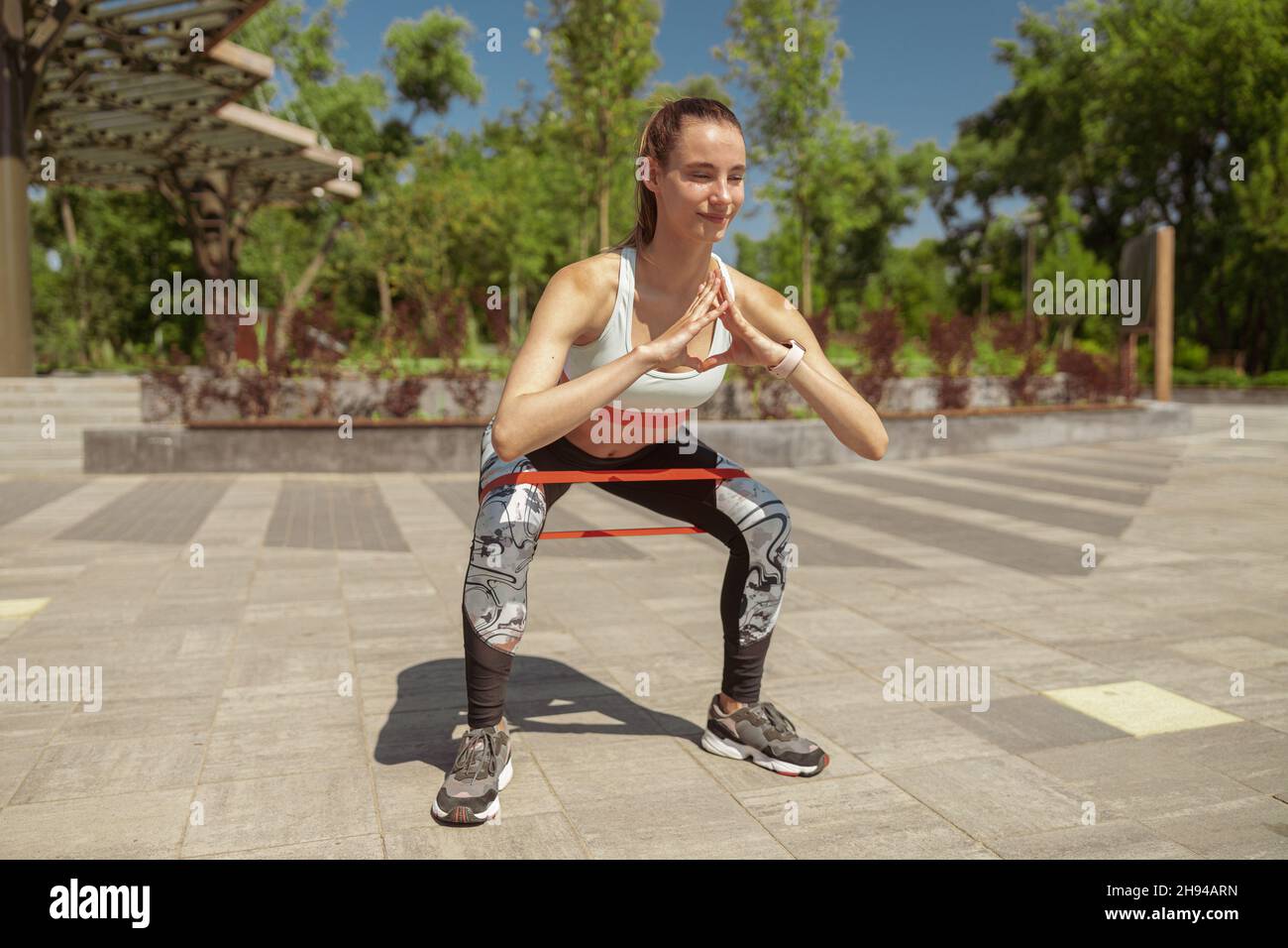 Smiling lady in sports clothes does squats with elastic bend in park Stock Photo